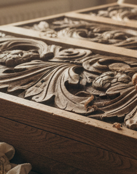 A cozy restoration workshop scene with a close-up view of hands carefully working on an ornate antique wood furniture piece, focusing on detailed carving and fine finishing tools on the bench. No face or head visible in the frame, just the torso, arms, hands, and the intricate antique piece in a warm, professional shop setting.